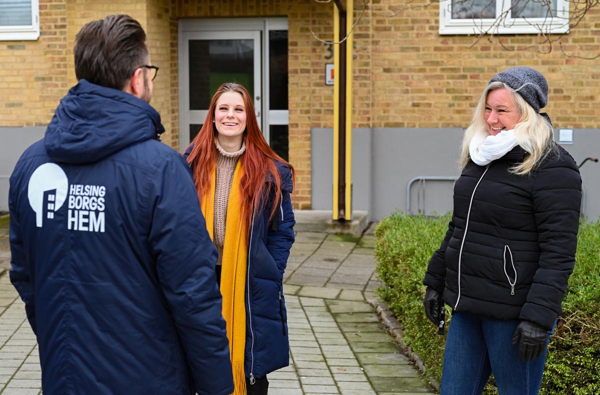 Två kvinnor och en man står framför ett bostadshus i Helsingborg. Mannen har Helsingborgshems logotyp på ryggen av jackan.